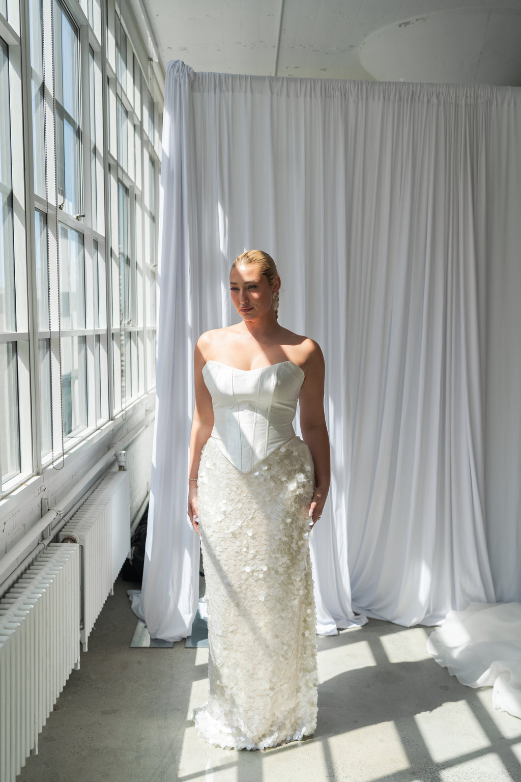 A woman wearing a strapless white wedding gown with a structured corset-style bodice and a large textured sequin skirt, standing in a bright room with large windows and white drapes.