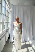A woman wearing a strapless white wedding gown with a structured corset-style bodice and a large textured sequin skirt, standing in a bright room with large windows and white drapes.