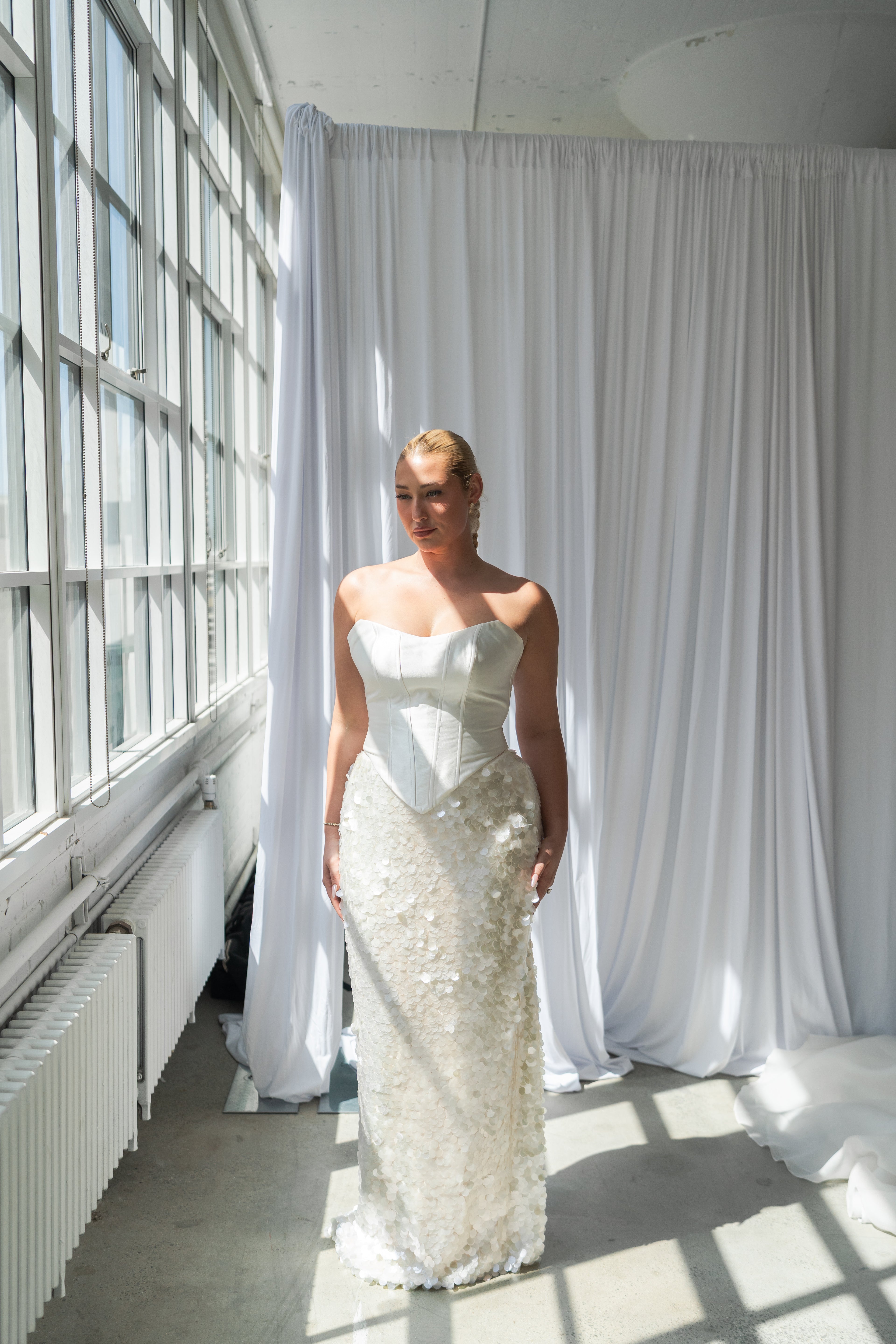 A woman wearing a strapless white wedding gown with a structured corset-style bodice and a large textured sequin skirt, standing in a bright room with large windows and white drapes.