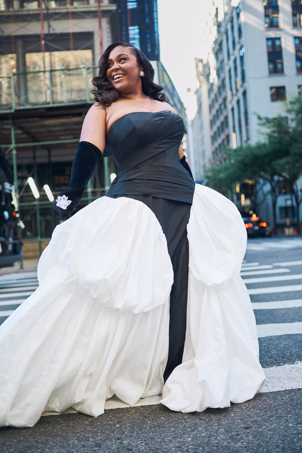 A woman modeling a dramatic black and white wedding gown in a ballgown style with long black gloves, standing on a city street with buildings in the background.